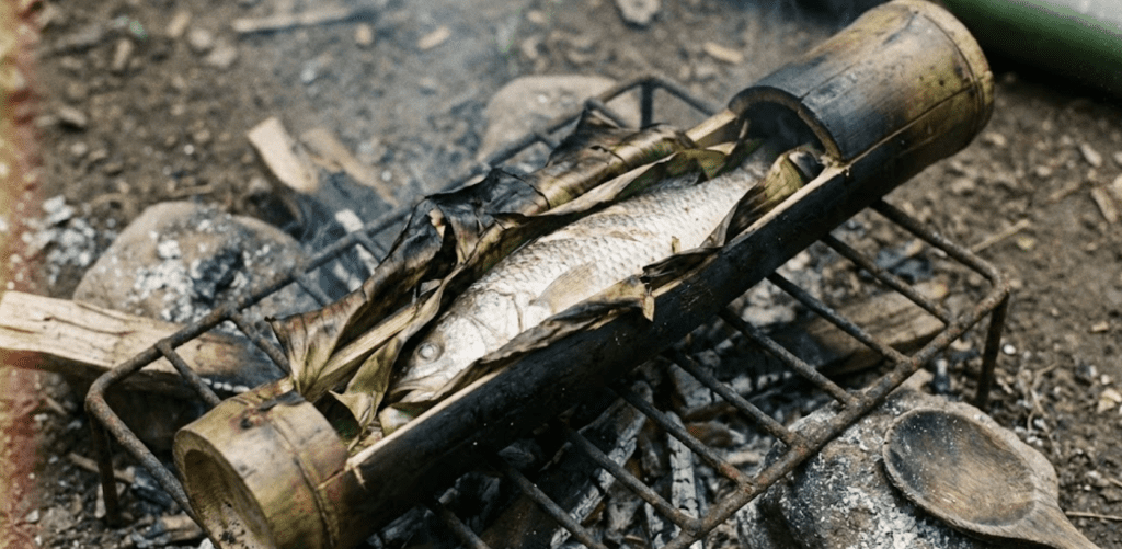 Fotografía realista de pescado cocinándose dentro de una tacuara (bambú) sobre brasas de leña en una cocina rústica de Pando, Bolivia.